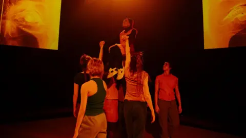 Luca Truffarelli A group of dancers wearing short sleeve tops and jogging bottoms standing together holding up an older woman. The woman is wearing black and orange clothing and white trainers, holding onto the dancers hands. The background is dark with two large orange pictures.