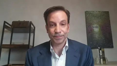 Erik Slavin wearing a white shirt and blue blazer, a grey wall in the background with dark metal-framed shelves with objects on display to his right, and a textured, greenish relief object on a thin stand to his left.
