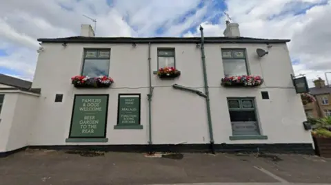Google Front view of The Milton Arms pub, a white-painted two-storey building with green trim, flower baskets beneath the windows, and signs advertising food, real ales and a beer garden.