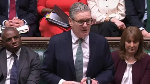 UK Parliament/PA A man, Keir Starmer, stood in the House of Commons. He is flanked by a woman, Rachael Reeves, to his right, and David Lammy, to his left, who are both sat on a green bench.