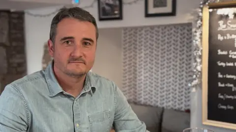 White man, Wesley Birch, with a brown hair and a close bead wears a blue denim shirt and looks directly at the camera. He's sat in a pub and a blackboard, bench and Christmas decorations are viable in the background. 