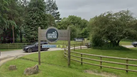 Cars leaving a surfaced drive way in a green space. A sign reads "Cotswold Farm Park". 