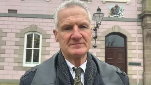Tom Binet. He has short grey hair and is wearing a jacket, a tweed jacket underneath and a white shirt and dark tie. He is standing in front of a government building.