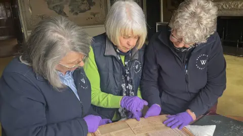 Three elderly women wearing Wentworth Woodhouse uniforms are examining an old document on yellowed paper. One woman points to it in a purple glove.