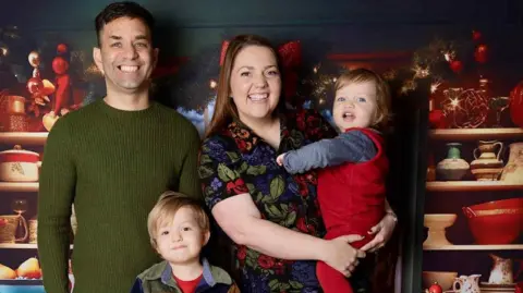 Zoe Rhiane Photography A man with dark hair grins at the camera, wearing a green jumper. A young boy is standing in front of him. Next to the man a woman stands, holding a small child. All are smiling ahead as they pose. Shelves are behind them with Christmas lights and kitchenware.