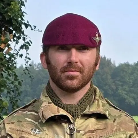 Ministry of Defence A soldier with a ginger beard dressed in his camouflage uniform, wearing a red beret, with a paratrooper emblem. He stands against a backdrop of green trees in the distance.
