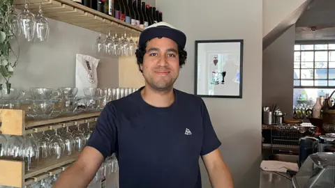 A man stands behind a café counter, with shelves of glassware and wine bottles, hanging lights and food displays visible around the venue.