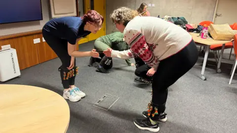 BBC/ Emily Johnson Liese and Hetty, who wear ankle bells, lean forward and clap their hands together.