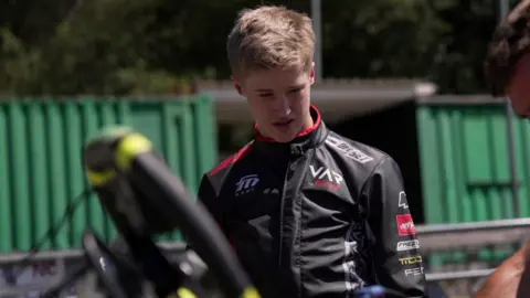 Noah in black racing overalls, watching a car being worked on. He has short light brown hair.