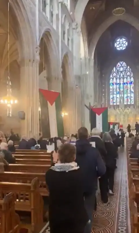 @SocialRightsIRL A place of people walked down the nave of a church with pews on either side. Some of the people are holding placards with Palestinian flags. In the background a large stained glass window.