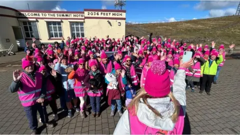 A woman hold a phone up to a crowd of people waving and wearing pink.