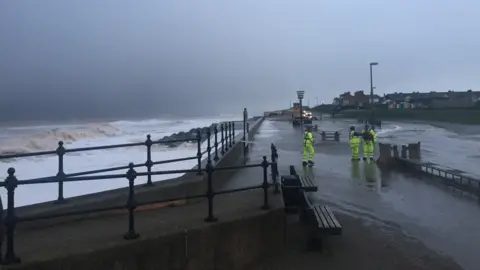 Sea wall and metal rails and rough sea on one side and three people in fluorescent stood on a wet promenade.