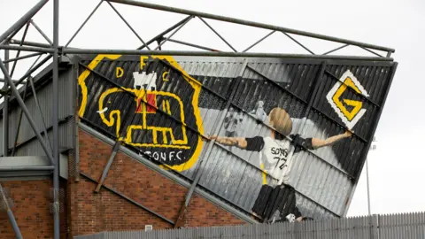 The outside of the Dumbarton Football Stadium, known as the Marbill Coaches Stadium. The stand is made of brick with a metal side and roof. On the side, a large mural of a player wearing a white shirt with black sleeves and yellow trim and the print 'sons 72' on the back has his arms outstretched in celebration. A large mural of the club crest, which is black with a yellow trim and a large black elephant with a yellow outline is in the upper left corner.