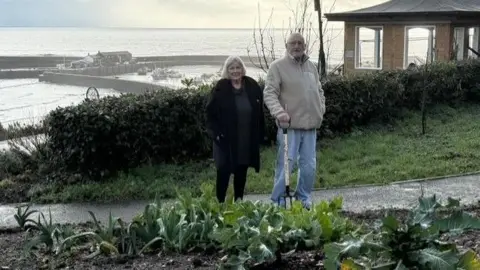 Cheryl and Alan Reynolds standing on a path at the community gardens in Lyme Regis with the harbour in the background.