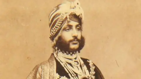 A portrait of Duleep Singh on paper that has aged over time and gone brown. He looks away from the camera and is expressionless. He has a 19th Century traditional Indian turban on as well as clothing. He also has a dark and thick beard.