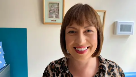 Ollie Conopo/BBC Jane Lafferty with brown bob-style hair cut, smiling in a leopard print top. She is in front of a white wall with a picture and digital clock on the wall.