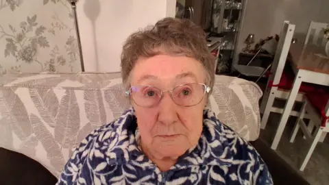 This is a photograph of Margaret Lister who lives in Eastfield in Scarborough. The photo shows her face and shoulders as she looks into the camera. Mrs Lister is wearing spectacles and a blue and white patterned top. She appears to have short brown hair which is greying at the sides. She is seated in a comfy chair in her living room. In the background is a wooden table with wood chairs tucked underneath. There is also a cabinet in the background which contains glassware.