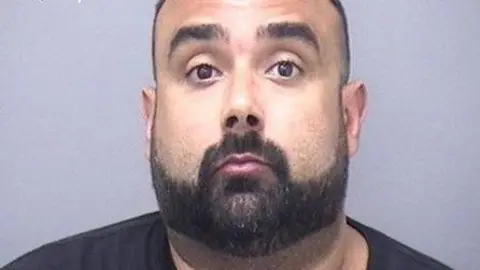 A mugshot of a bearded man with raise eyebrows looking directly into the camera lens, 