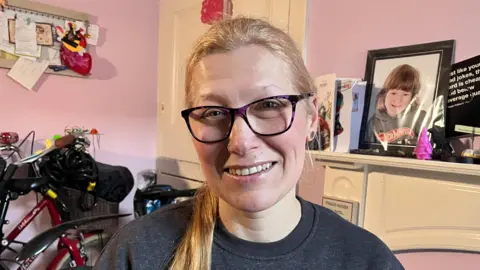 Monika sits in front of her fireplace and a photograph of her son and smiles at the camera wearing a grey jumper and dark purple glasses.