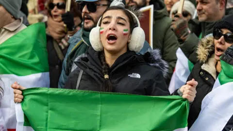 A woman wearing earmuffs and holding a flag shouts at a protest
