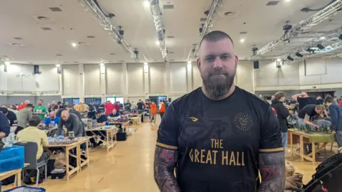 Liam Dempsey, a man with short hair and a beard, is stood in the hall. He's wearing a black sports shirt and has two sleeves of tattoos. 