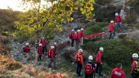 Mountain rescue team carrying a stretcher along a rocky hillside during a training exercise.
