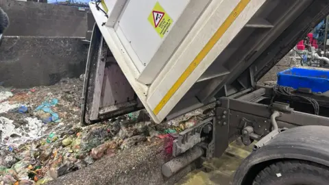 Kate Bradbrook/BBC Inside the facility, a truck tips backwards to release lots of plastic bags of food waste. You can see some orange food amongst it all. 