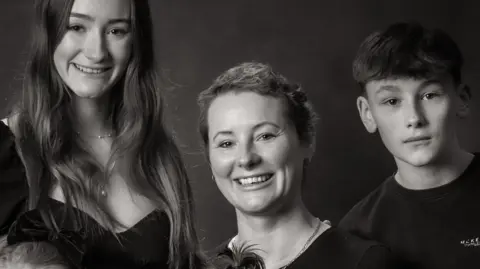 Supplied A professionally-taken, black and white photograph showing a woman with short hair sitting down. On her lap, is an infant girl. Next to her are her two daughters, one aged 19, the other five, and her teenager son.