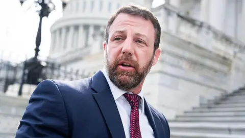 Markwayne Mullin wears a navy blue suit and a red patterned tie. In the background are white steps and the Capitol building in the distance. 