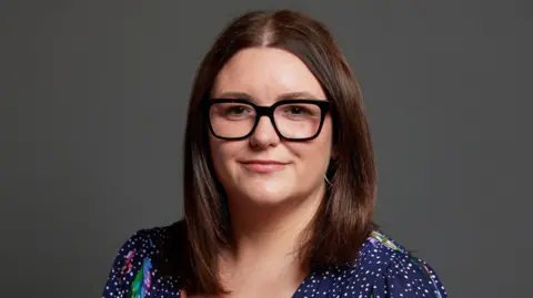 UK Parliament Leigh Ingham, a woman wearing a navy blue dress with white spots and a coloured pattern. She is pictured against a dark grey backdrop.
