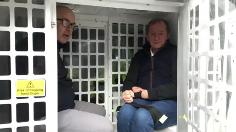 Andy Cocksedge wearing a black coat and grey trousers sits in the back of a police van next to Michael O'Hagan wearing a dark green jacket and jeans. His legs tied together with a black strap and his hands are bound by handcuffs. 