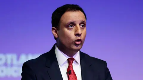 Anas Sarwar wears a dark suit with white shirt and red tie. He has short dark hair and stands in front of a purple screen