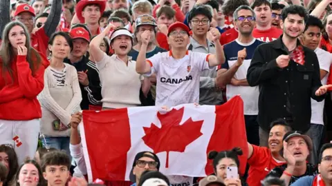 Reuters A crowd of Canada fans, many in red and white, react to a play by Canada. Two are holding up a Canadian flag. 