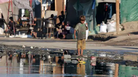 Getty Images Seorang anak laki-laki Palestina berdiri tanpa alas kaki di dekat genangan air limbah di Deir el-Balah, di Jalur Gaza bagian tengah (19 Juli 2024)