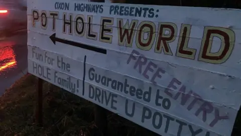 A sign reading: "Oxon Highways presents: Pothole World", "fun for the hole family" and "guaranteed to drive you potty". It is right next to a road.