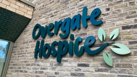 The outside of the new Overgate Hospice. The building's logo is written in dark green letters with a light green flower emerging from the final 'e'. It is on a brick wall background.
