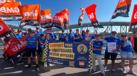 Pippa Unwin/LDRS A number of people in blue t-shirts hold orange GMB Union flags and a large banner which reads 'support your coastguard rescue officers'