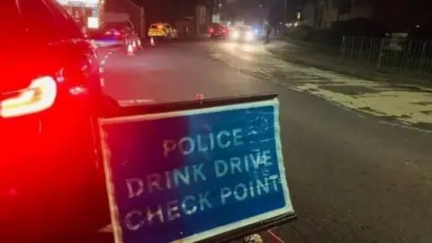 Cambridgeshire Police A blue and white police sign that reads "Police Drink Drive Check Point", stationed behind a line of queueing cars. 