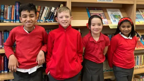 A mixed gender group of Fishergate Primary School pupils stand in front of wooden library bookshelves. They wear a mix of school uniforms; red jumpers, polo shirts and fleeces, with grey trousers. 