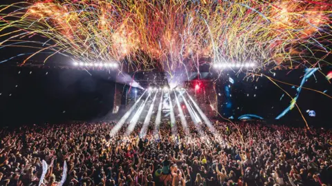 A large crowd of people are watching a performance at Creamfields as streamers fall from above and rays of white light are projected from the stage. 