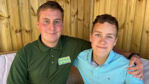 The two brothers smile into the camera. Harry is wearing a dark green polo with a label on the left side reading 'Aston Project'. Arthur is wearing a light green polo. Harry has his arm around Arthur. 
