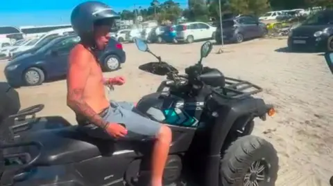 Family handout Alfie with no top on in the sunshine on a quad bike, wearing a helmet