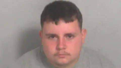 A custody photo of a man wearing a grey jumper. He is looking at the camera and has short brown hair.
