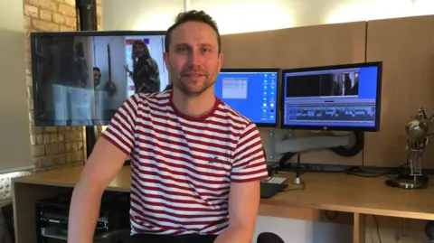 Fran Ackroyd Steve Ackroyd is seen wearing a red and white striped t-shirt sits in a video editing suite, surrounded by monitors displaying editing software and footage.