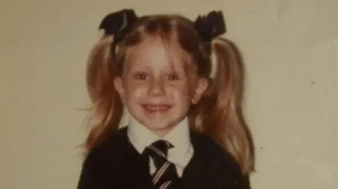 SWNS Lizzie as a child. She had blonde hair tied in pigtails with ribbons and is wearing a school uniform