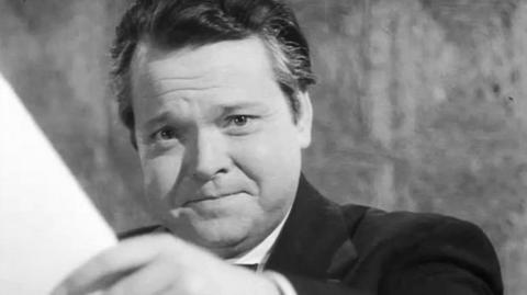 Orson Welles holds a piece of paper in front of the camera.