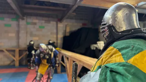 A man in a helmet and a colourful tunic looks into the distance. He's leaning on a wooden barrier with other armoured men in the background.