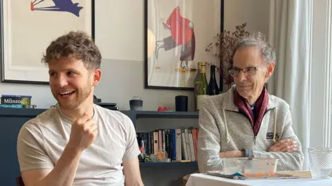 Jade Lau David has light brown hair and wears a white t-shirt and looks off to the side. He is sitting in a living room setting with framed prints behind him. He sits next to his dad, Richard, who wears a three quarter zip jumper and glasses. He smiles while looking in the same direction as David.