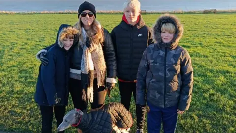 Family handout Three boys with winter coats, two are dark, one is camouflage patterned. Two have their hoods up. They are all wearing dark trousers and trainers and have short blonde hair. A lady with a hat, sunglasses and scarf also has a winter coat, dark trousers and boots. There is a lurcher dog with them with a winter coat. They are standing on a pavement with grass, water and a clear sky.