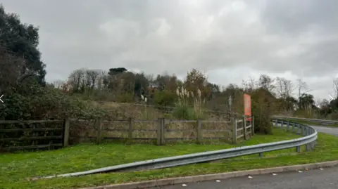 A grassy area near a road. On the right is the entrance to Gables in Plymouth. There is also a wooden fence which has bushes and trees behind it.